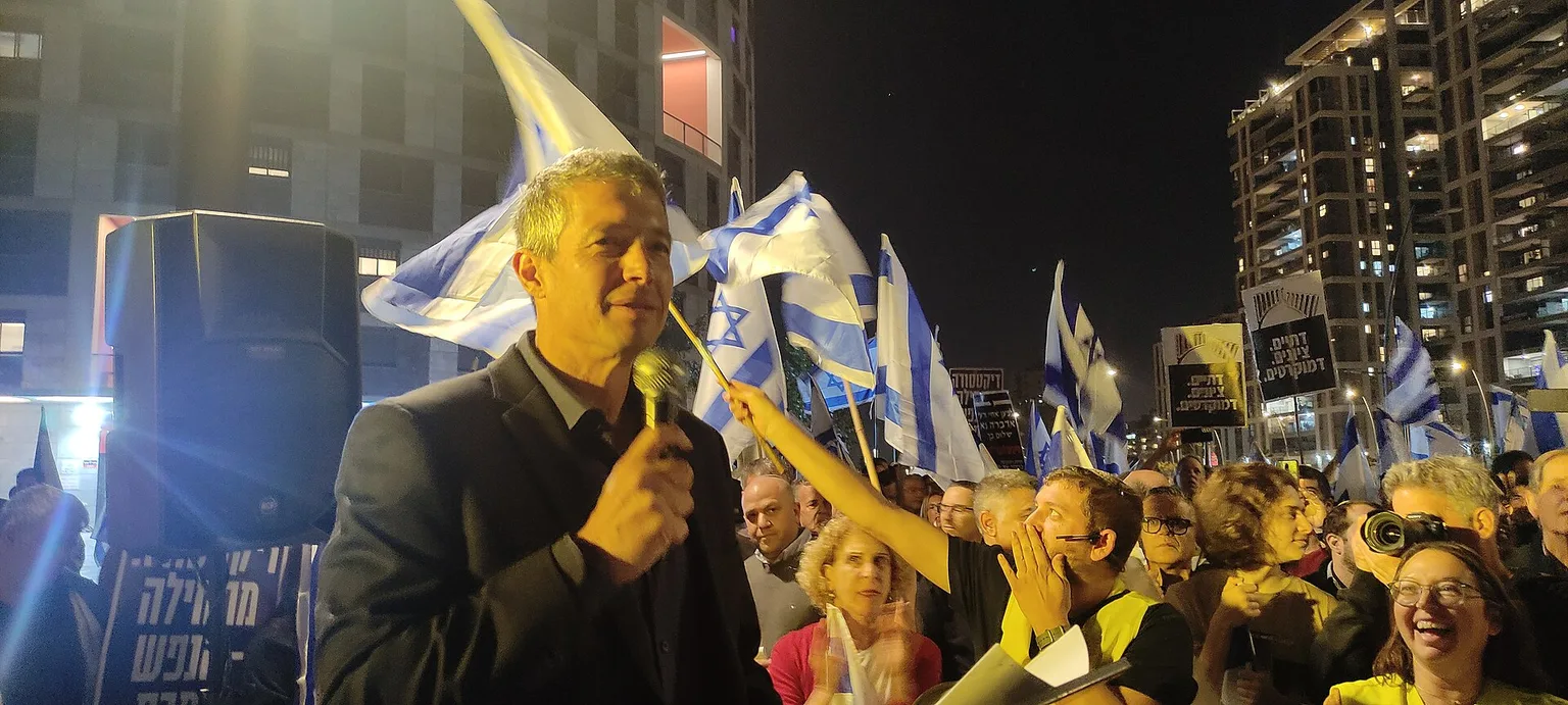 יועז הנדל נואם בהפגנה, כשברקע מפגינים ודגלי ישראל