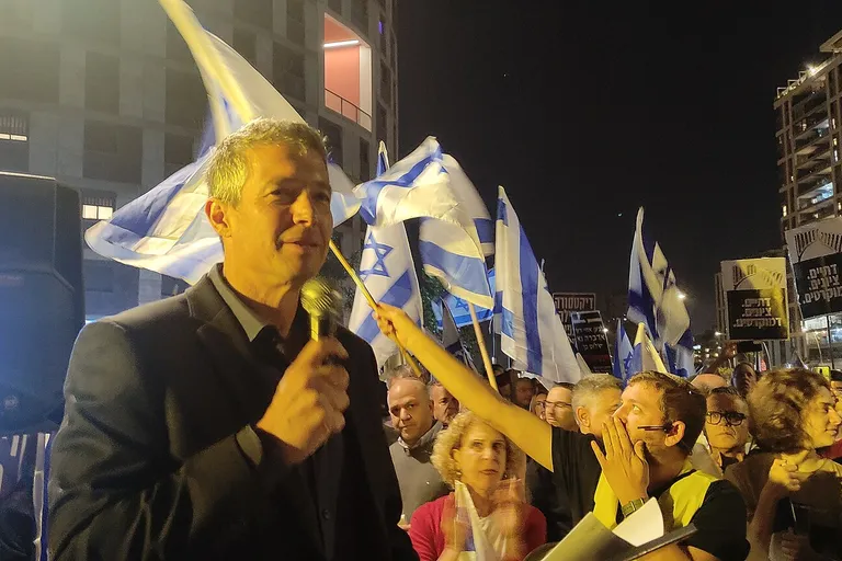 יועז הנדל נואם בהפגנה, כשברקע מפגינים ודגלי ישראל